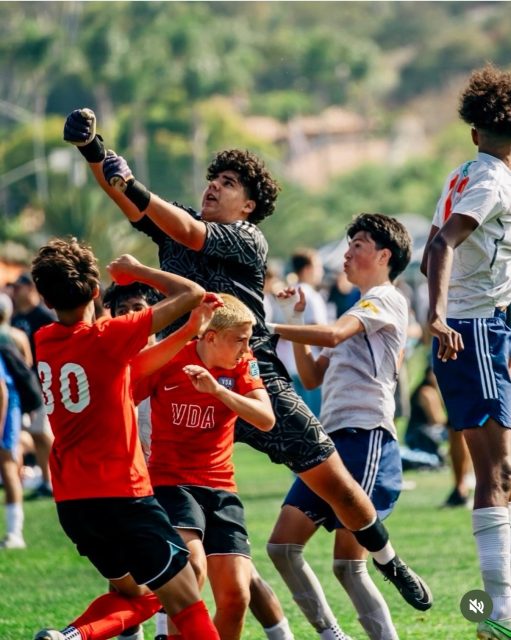 A soccer goalie jumps to punch the ball away as players in red and white jerseys compete during a match on a grass field.