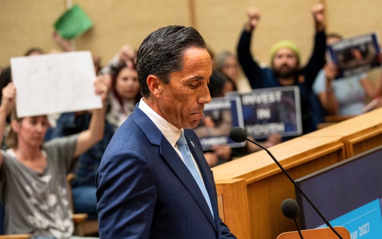 Todd Gloria on his phone with people holding thumbs down behind him