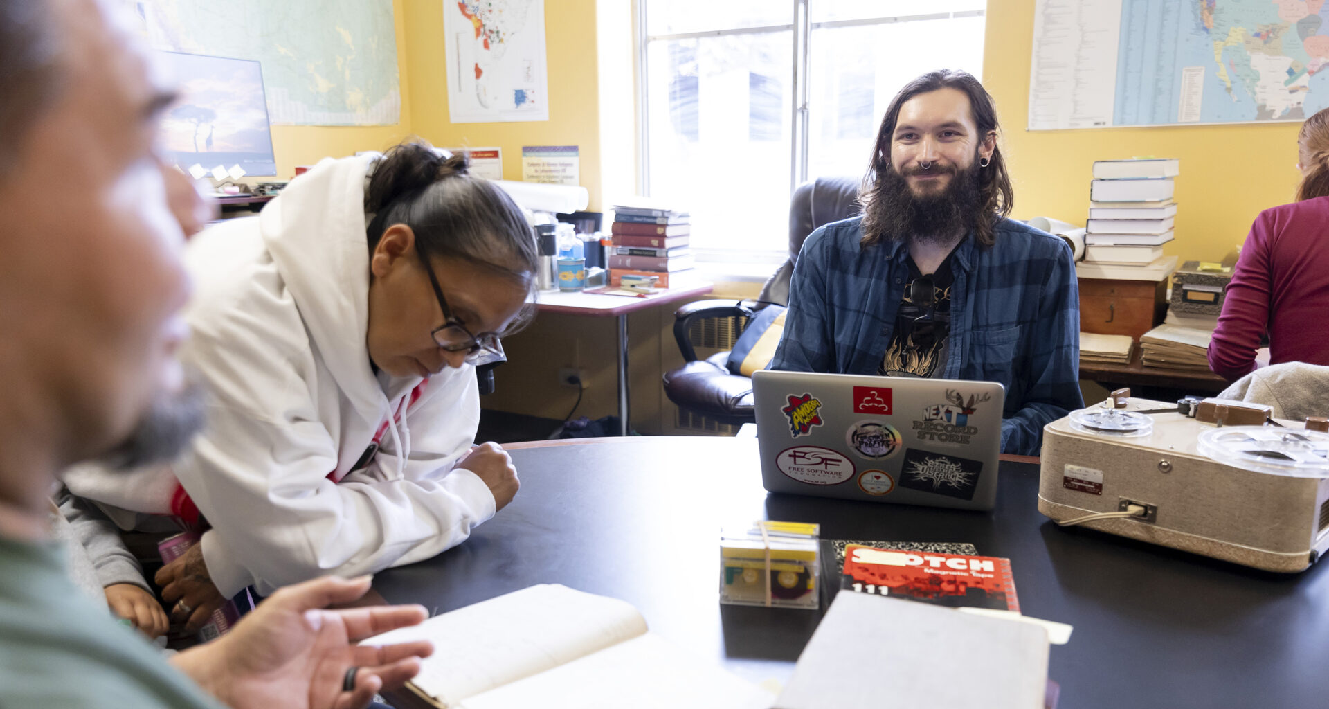Berkeley graduate student Tyler Lee-Wynant sits smiling at a table inside the California Language Archive, surrounded by archival materials, a laptop, an audio recording player while speaking with tribal visitors.