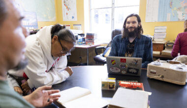 Berkeley graduate student Tyler Lee-Wynant sits smiling at a table inside the California Language Archive, surrounded by archival materials, a laptop, an audio recording player while speaking with tribal visitors.