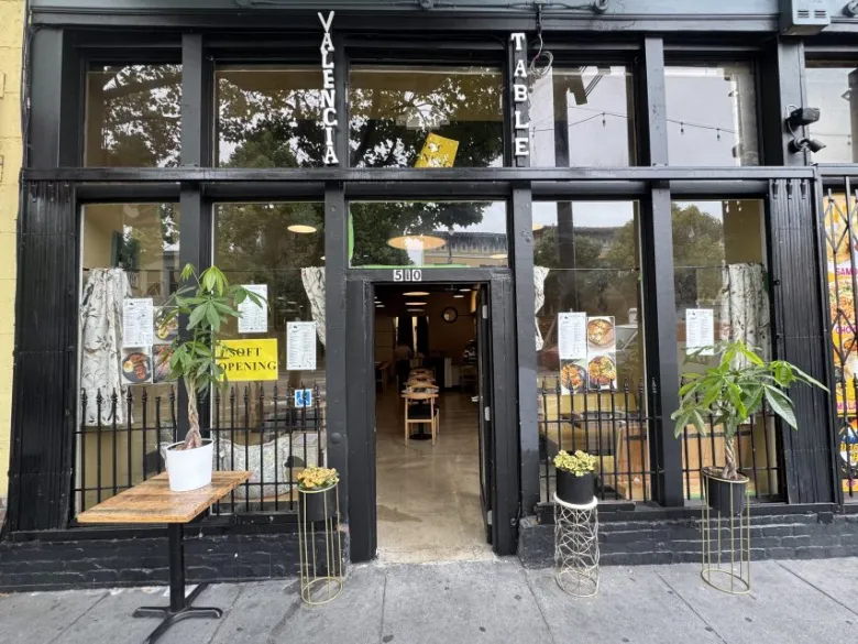 Street view of a restaurant with large windows, an open glass door, plants on tables and stands outside, and "Valencia Table" signage above the entrance.