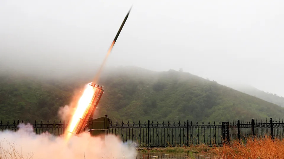 A rocket blasts off from a launch page next to a fence and forested hill.
