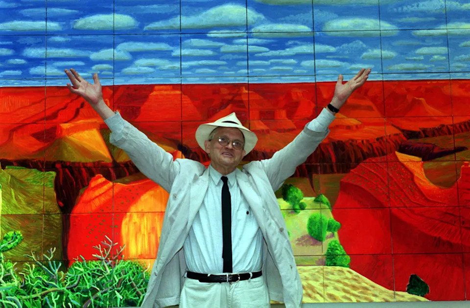 David Hockney standing with his arms outstretched in front of his artwork Grand Canyon II.