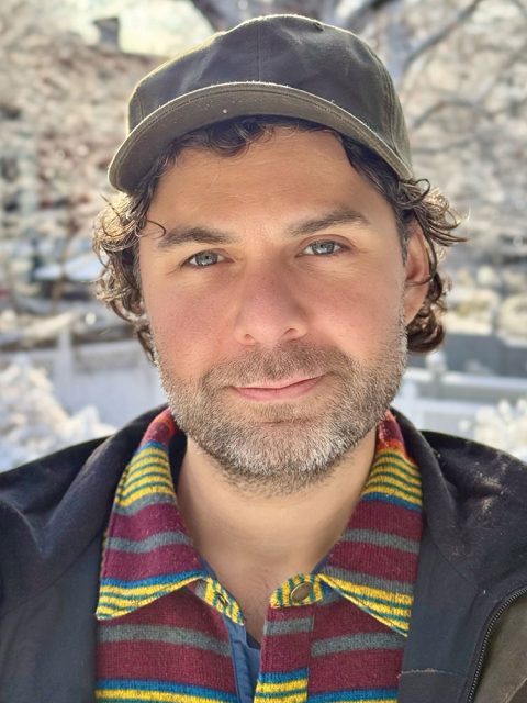 A man with curly hair, a short beard, and a green cap looks at the camera outdoors, wearing a colorful striped scarf and a jacket.