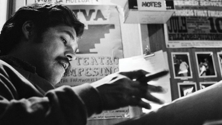 A man with a mustache writes or gestures with a pen at a desk, with posters and notes visible on the wall behind him.