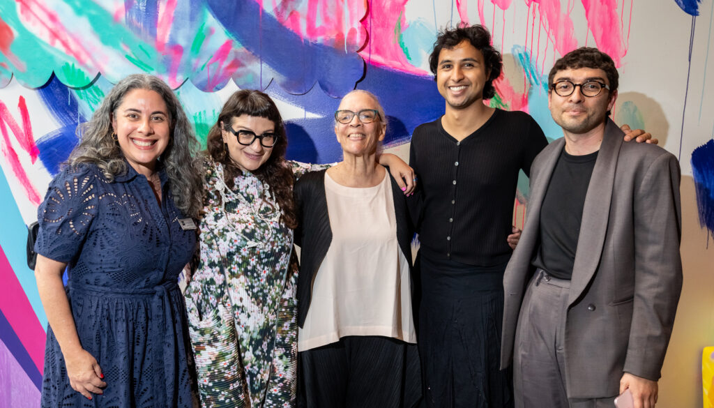 Artist Maren Hassinger stands in the center of a group of five BAMPFA colleagues, smiling arm-in-arm against a vibrant, multi-colored gallery wall.