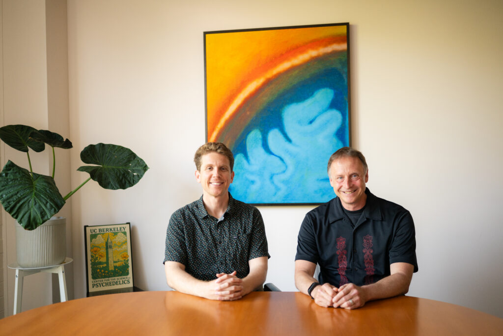 Two people sit at a table smiling at the camera. Behind them is a painting with blue swirls that are reminiscent of brain matter.