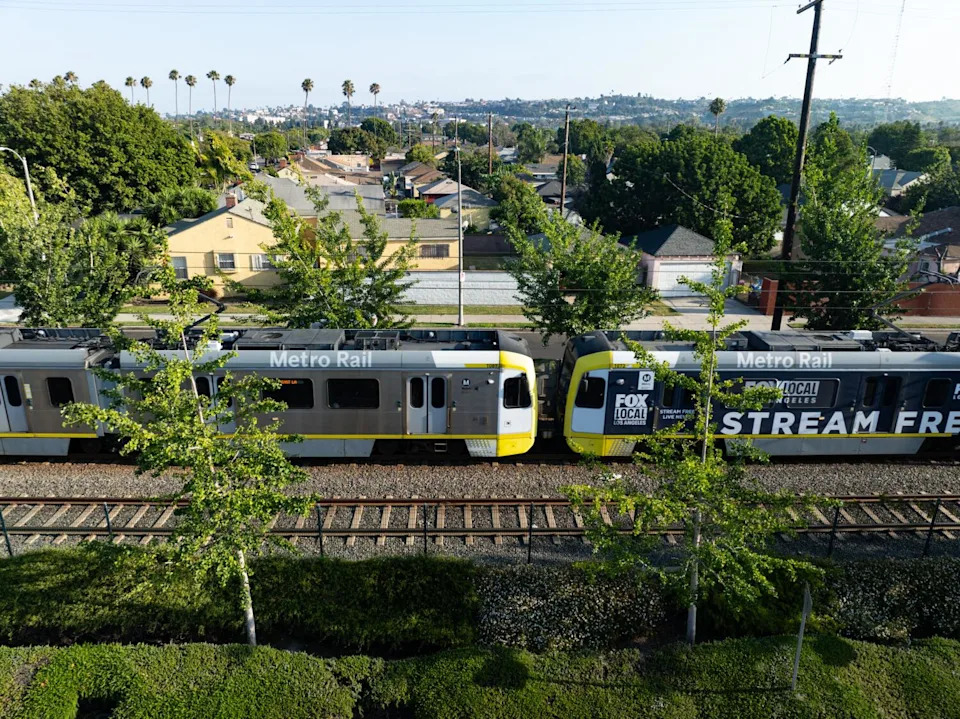 A light-rail metro train in Los Angeles passes through a neighborhood with houses and trees.