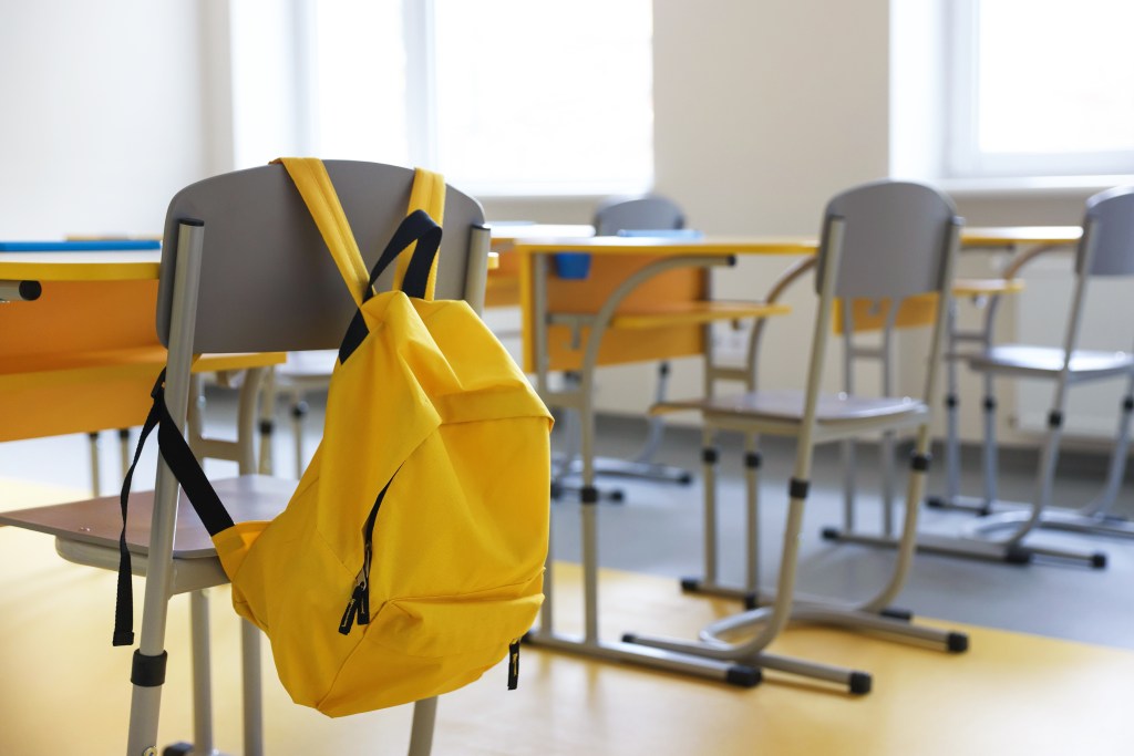 A bright yellow backpack hangs on the back of a grey school chair in a classroom with yellow desks and additional chairs.