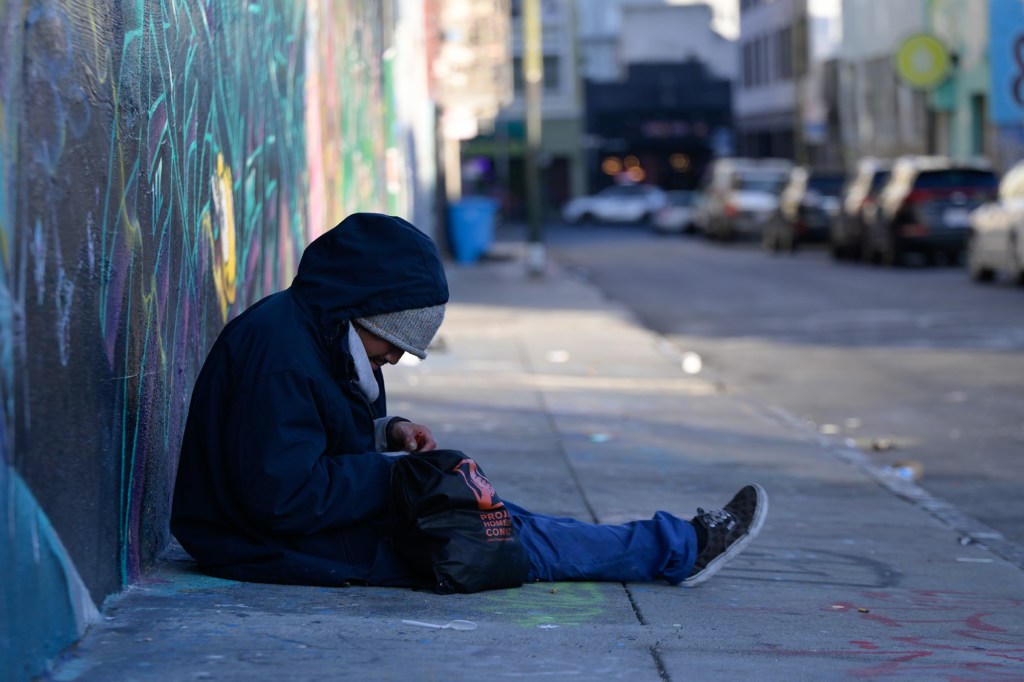 A person in a hooded coat sits on the ground next to a graffiti-covered wall, looking into a black bag.