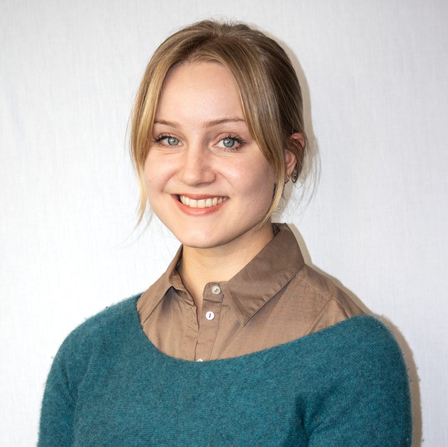 A woman with blonde hair, wearing a teal sweater over a brown collared shirt, smiles at the camera against a plain light background.