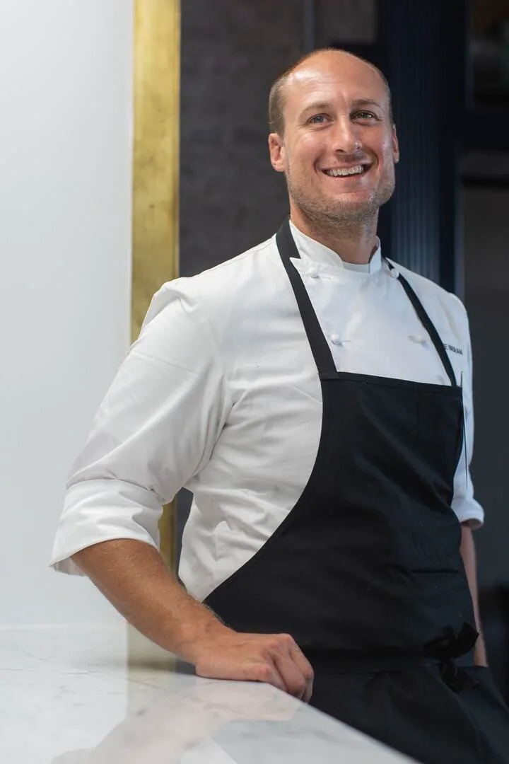 A smiling man in a white chef's jacket and black apron