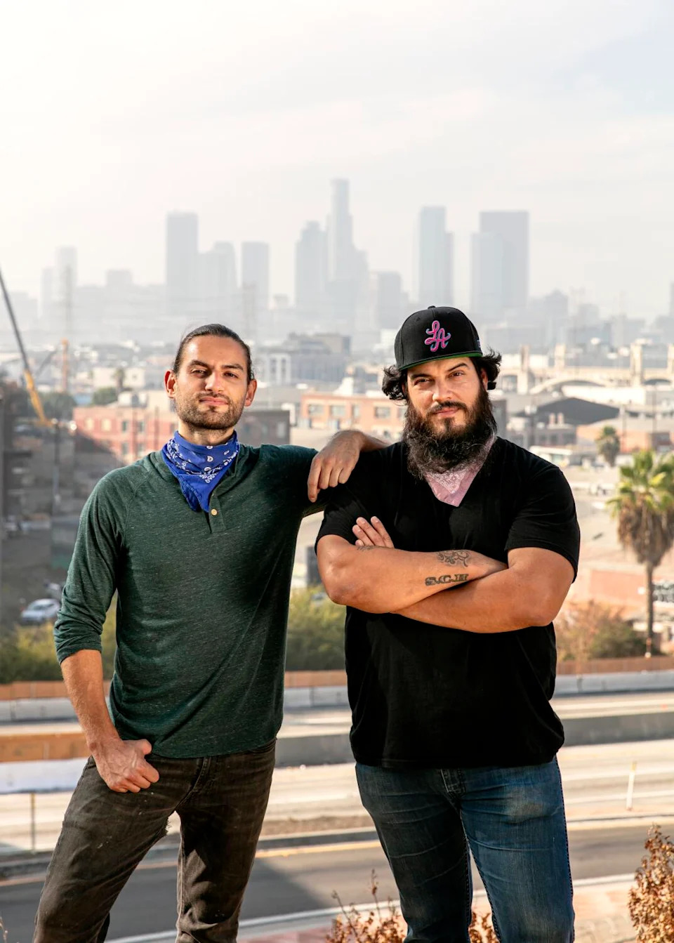 A man in a dark blue shirt standing next to another man, in a dark cap and short-sleeved shirt, arms crossed