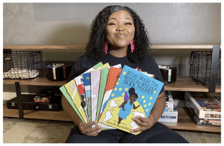 A woman sits on the floor, holding a stack of children's books, with shelves and board games visible in the background.