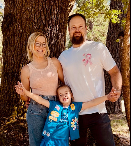 Woman with blonde hair and man with brown hair and beard stand in front of tree and hold hands with young girl in Girl Scout vest.