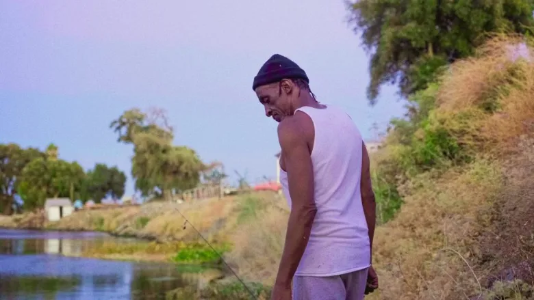 A person in a white tank top and black beanie stands on a grassy riverbank, looking down, with water and trees in the background.