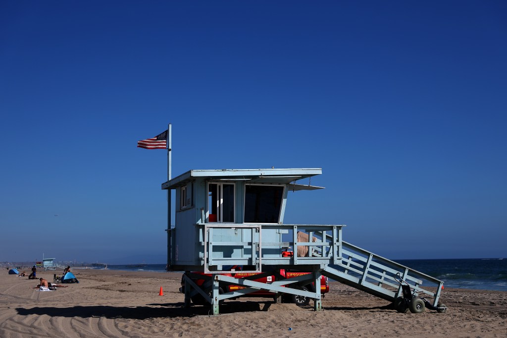 A standard light blue lifeguard tower with an American flag flying above it sits on a sunny beach, with the ocean in the background.