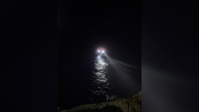 A rescue boat is on standby as first responders swim to a stranded victim on a cliff.