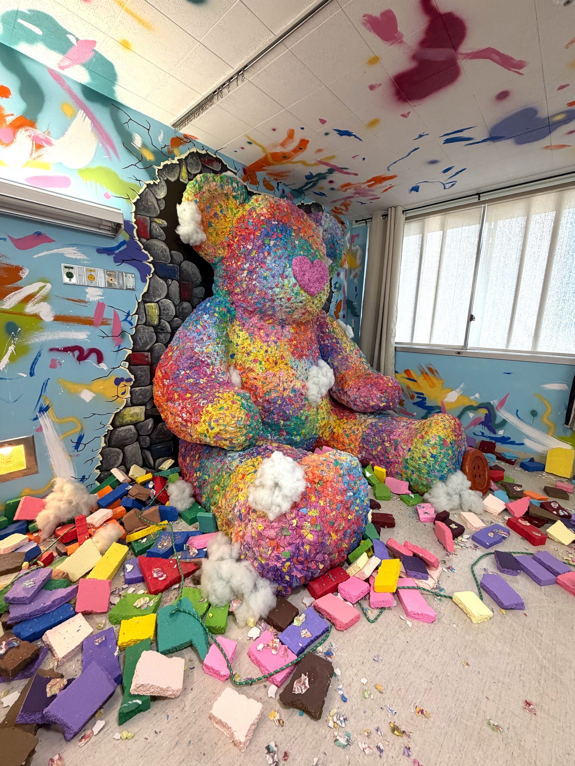 a hospital room installation transformed by an enormous colorful teddy bear bursting through a wall