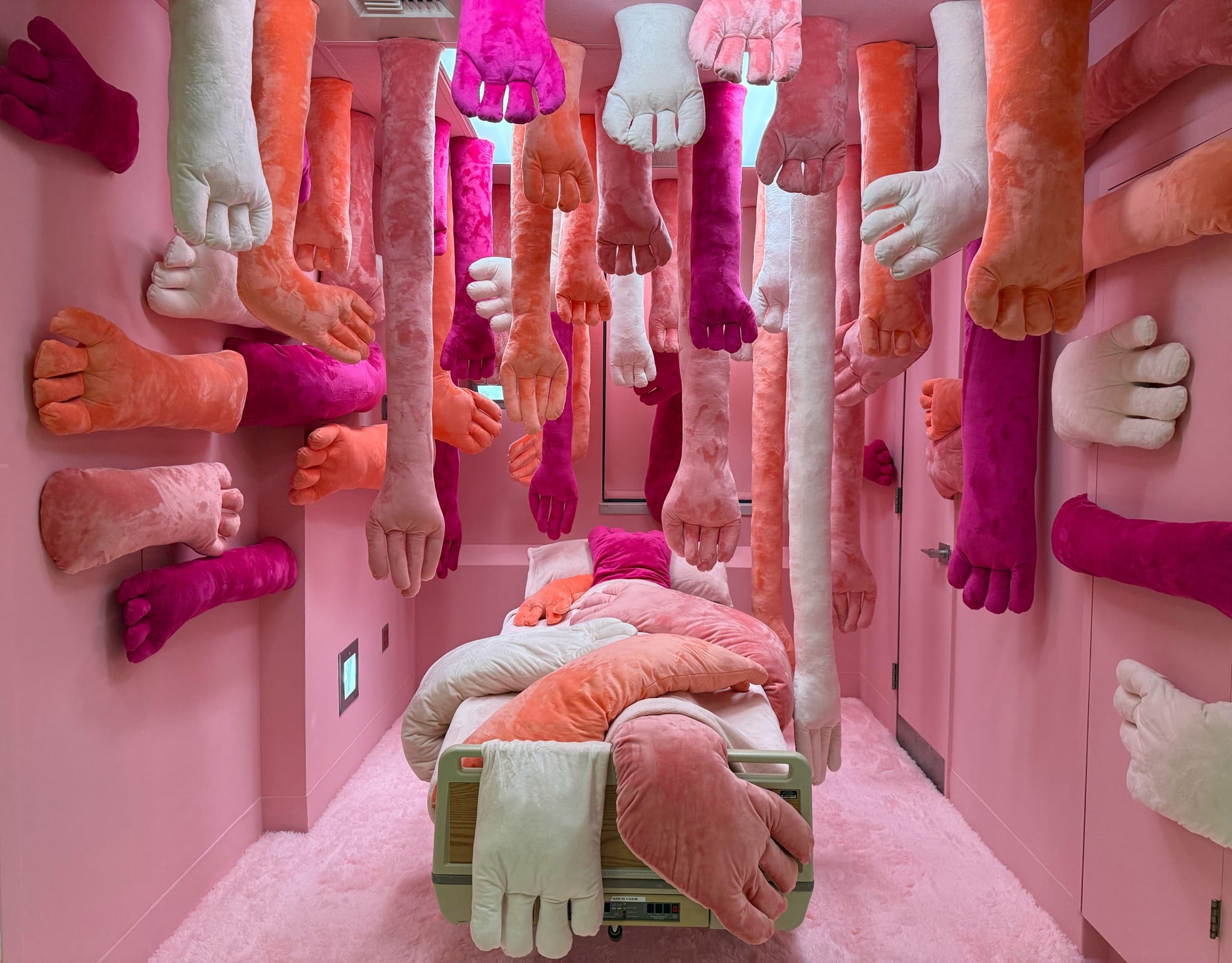 a hospital room installation transformed by pink walls and suspended plush arms and hands from the ceiling and on the bed