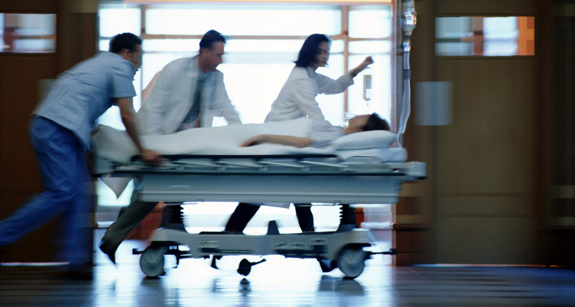a motion-blurred image of medical workers pushing a patient on a gurney down a hospital corridor