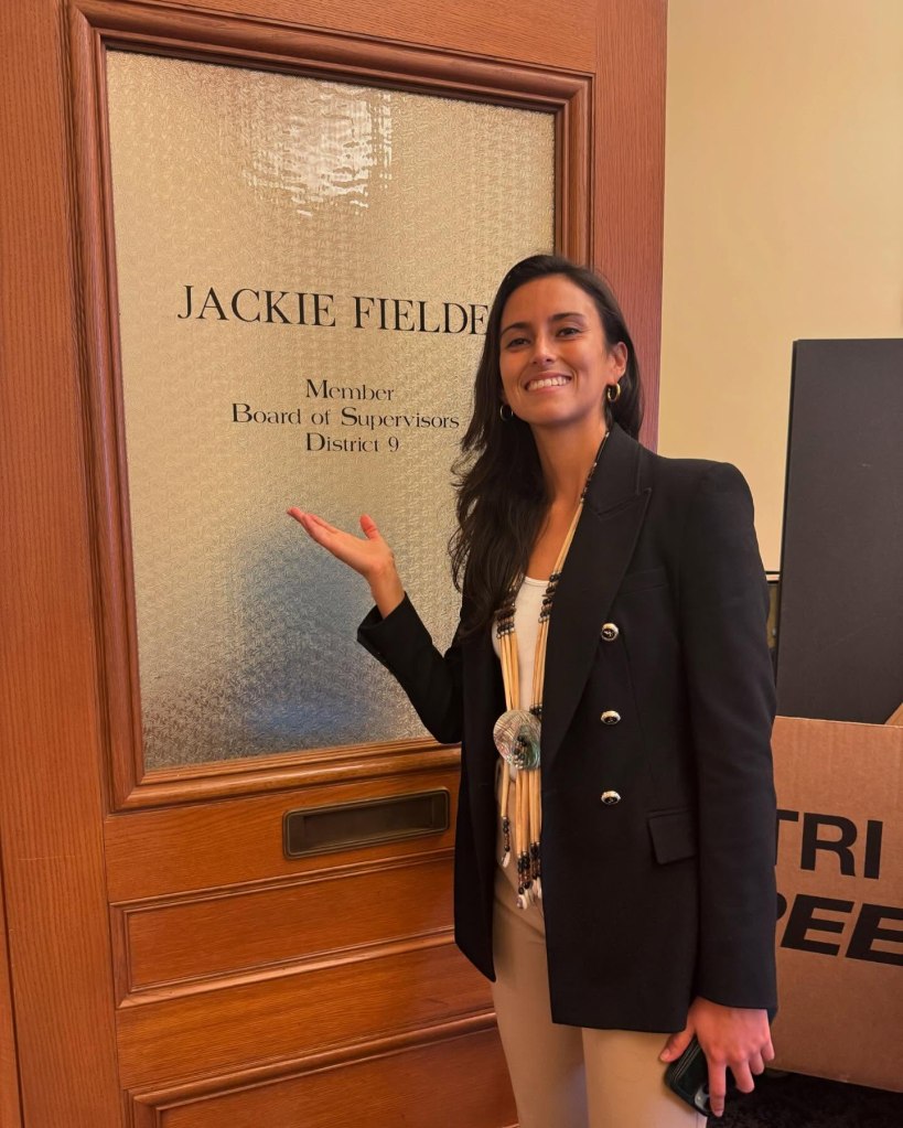 Jackie Fielder stands in front of a door with her name and title "Member Board of Supervisors District 9" on it.
