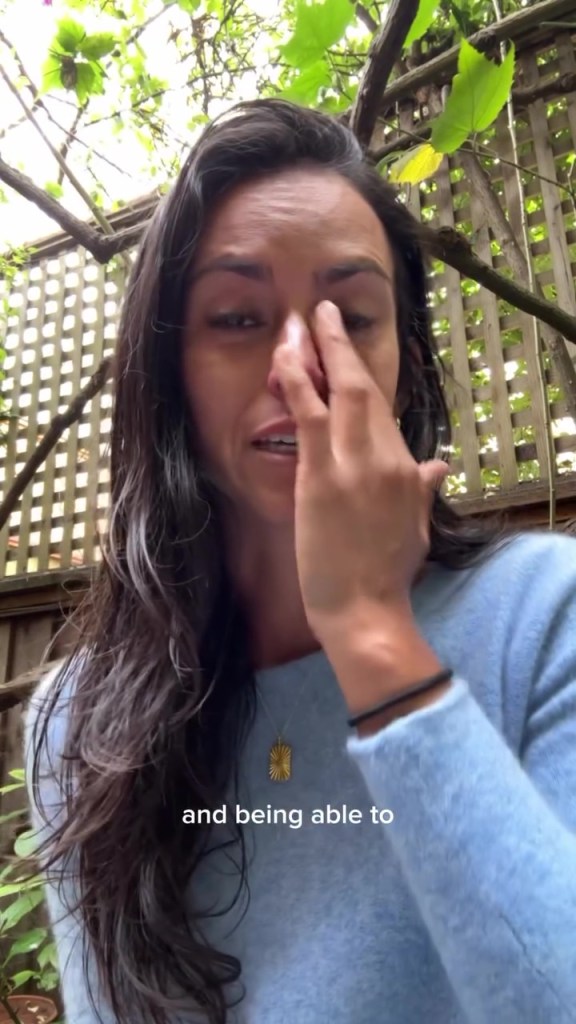 Jackie Fielder crying, dressed in a blue sweater with a gold necklace, outside with foliage and a lattice fence in the background.