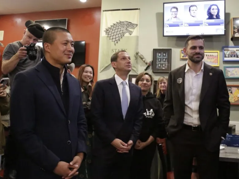 Three men in suits stand together at an indoor event, with people and photographers in the background and TV screens displaying election coverage above them.