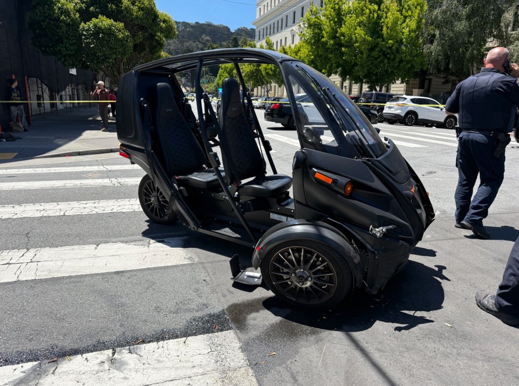 A black three-wheeled motor vehicle with an open top and two front seats, showing damage to the front right side, on a crosswalk at an intersection, with police and yellow tape in the background.