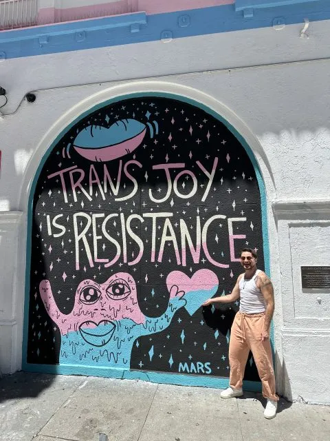 A person stands and smiles in front of a mural that reads "Trans Joy is Resistance" with colorful artwork including stars, a UFO, and a blue character holding a pink heart.