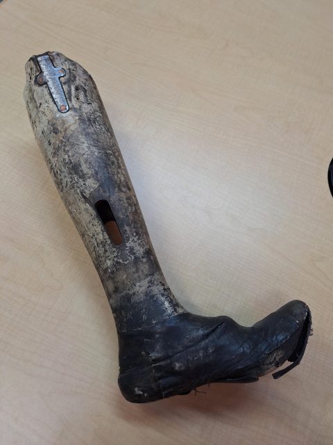 A worn, vintage prosthetic leg with a leather foot and aged, discolored surface rests on a light wood table.