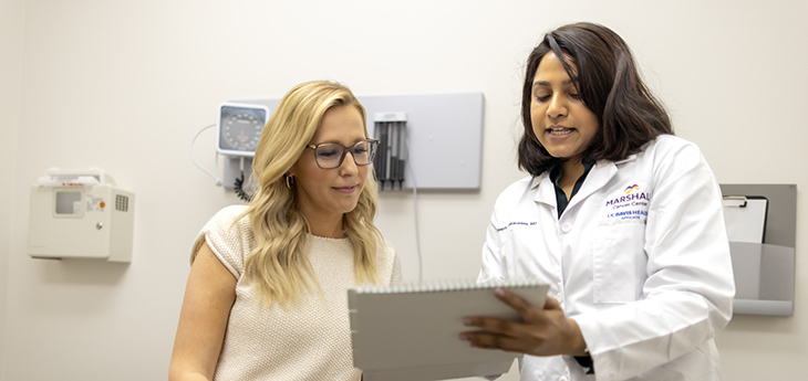 Blonde woman sits on patient exam table while woman with dark hair hold clipboard and speaks with her.