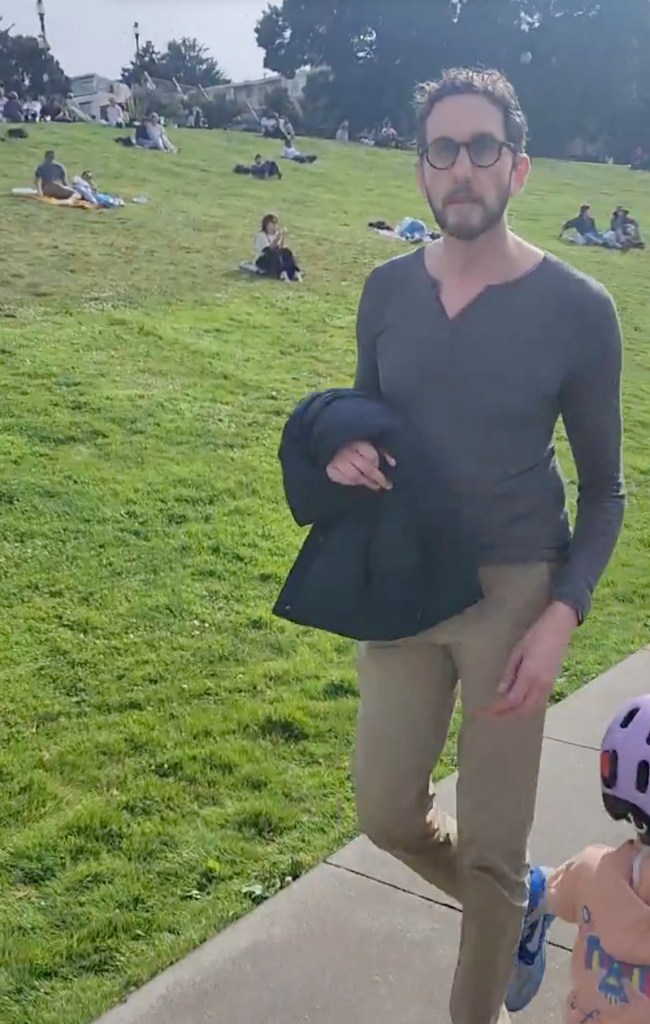 Scott Wiener, in a grey shirt and khaki pants, walking away with a jacket in hand from a group of people sitting on a grassy hill.