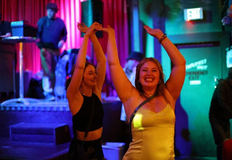 Two women are dancing with raised arms in a dimly lit venue with red curtains in the background. The woman on the right is wearing a yellow dress and smiling broadly.