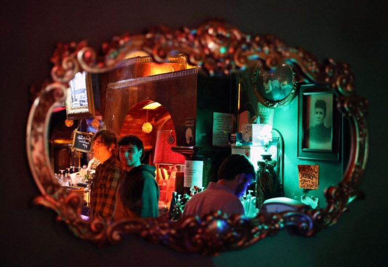 A decorative mirror on a wall reflects three people in a dimly lit bar, with colorful lighting and various posters on the wall.