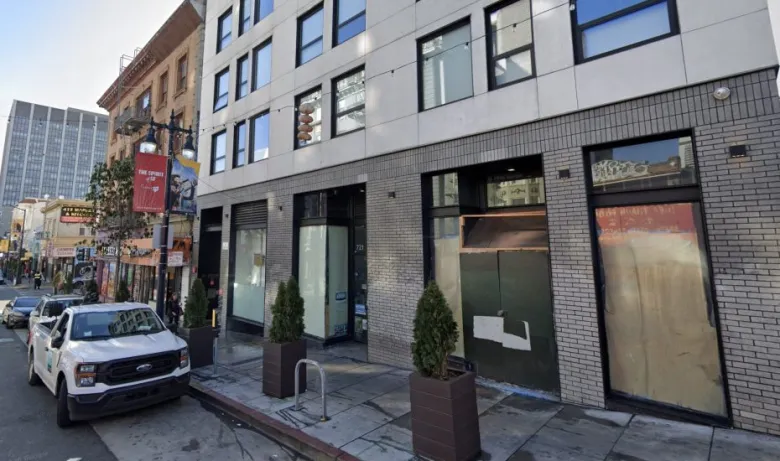 A city street with a parked white truck, several potted plants on the sidewalk, and a modern building with boarded-up and covered windows at street level.
