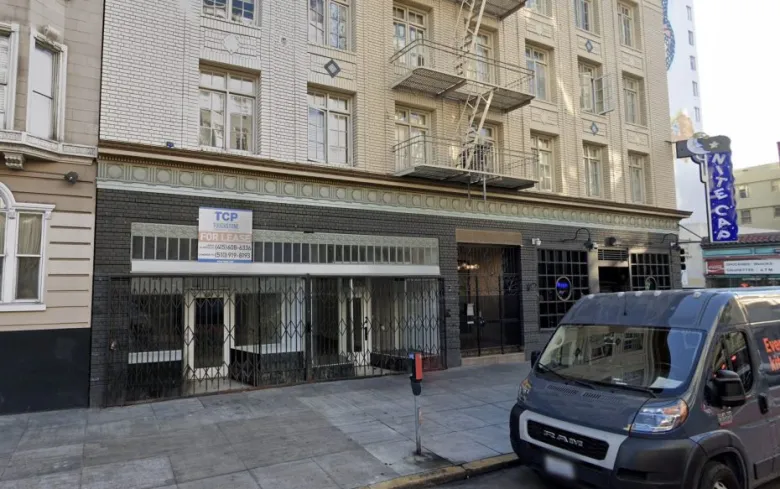 Street view of a multi-story building with a "For Lease" sign in the window, metal gates over the storefront, and a van parked at the curb.