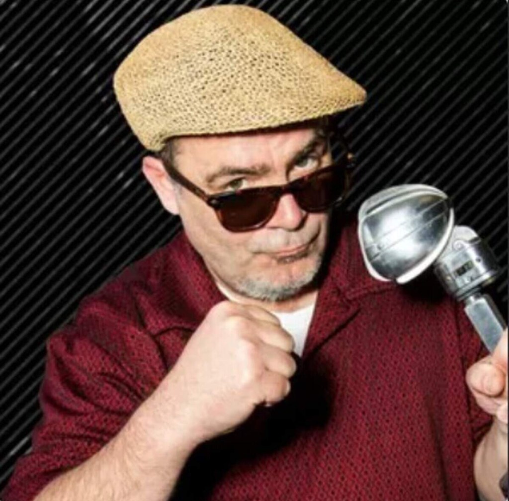 Trevor Carey, wearing a straw fedora and sunglasses, holding a vintage microphone and making a fist.