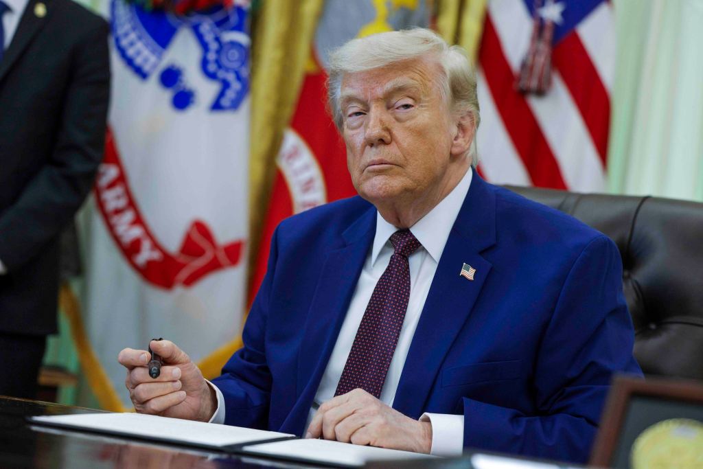 President Donald Trump in a blue suit and red tie, holding a pen, sitting at a desk during an executive order signing ceremony.