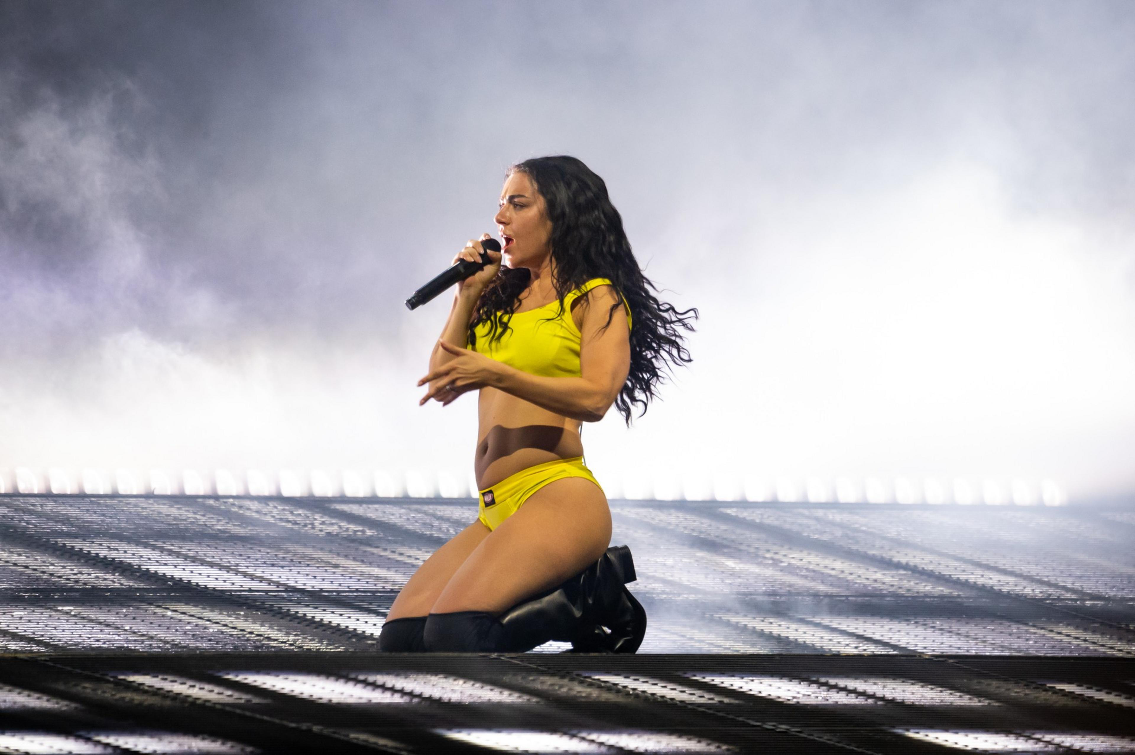 A female performer in a yellow outfit and black boots kneels on stage, singing into a microphone with a smoky, illuminated background.