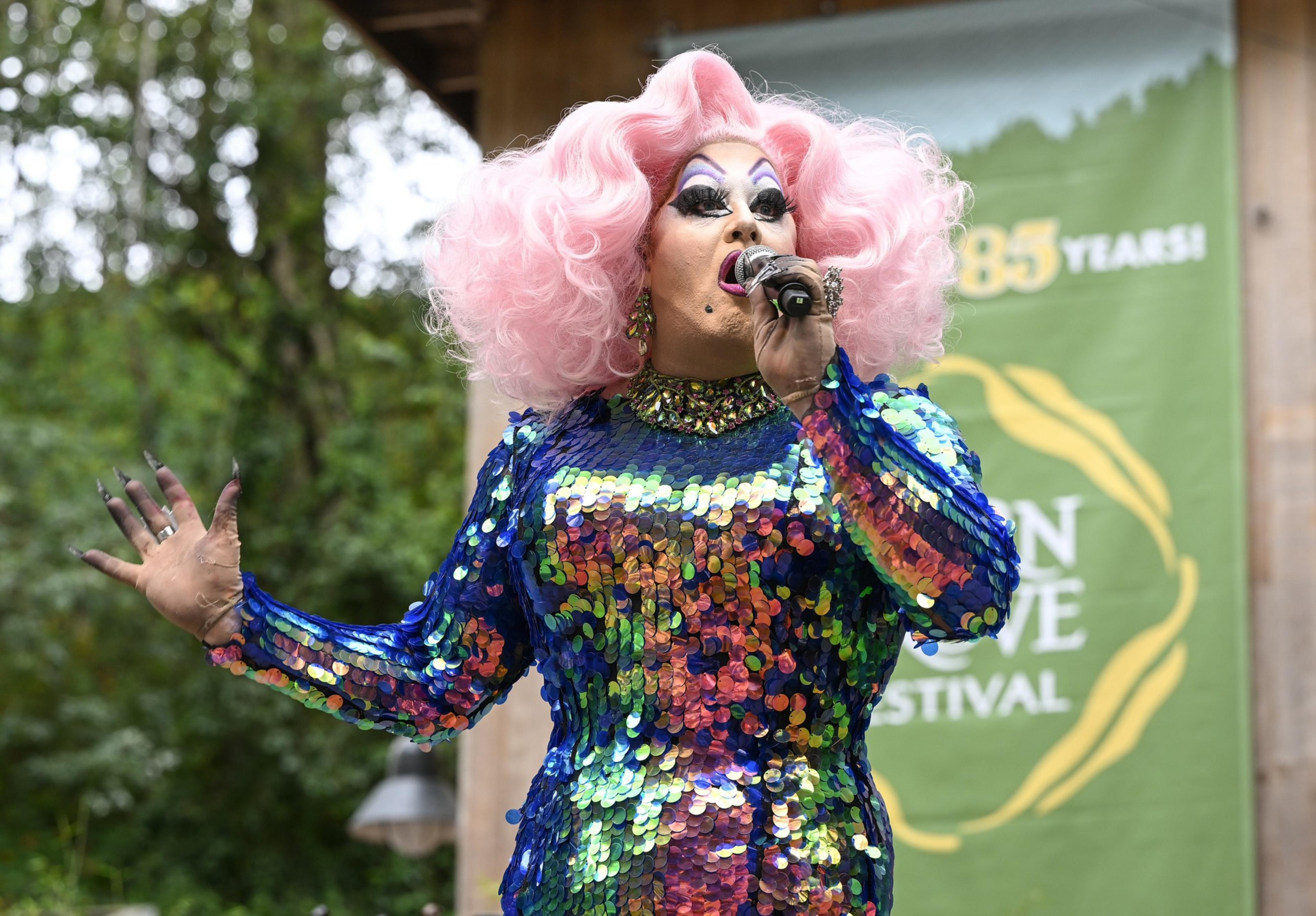 A performer with voluminous pink hair and dramatic makeup wears a shiny, multicolored sequined dress while singing into a microphone.