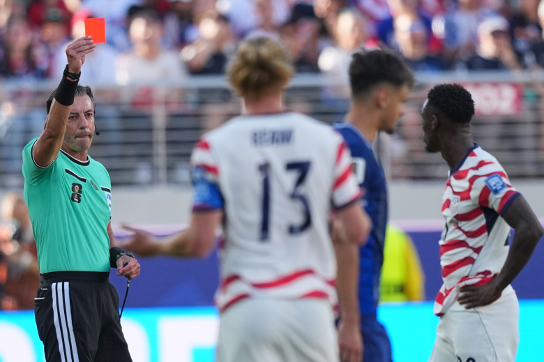 An official holds up a red card while one player passes by and two players stand near him, one in the center and one to the side. The one in the center has his arms out in front of him.