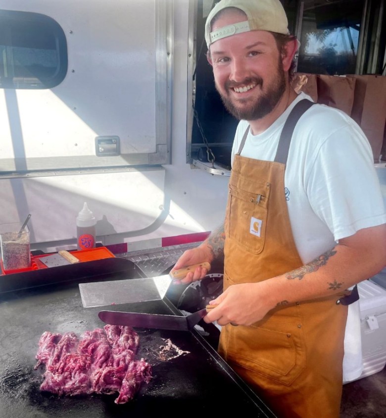 A man in an apron smiles into the camera while holding two spatulas over red meat on the grill.