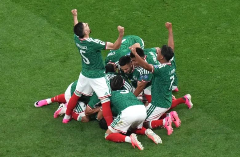 One man is on his knees on a soccer field, in a dogpile with several other men celebrating. One among them stands, bends back and raises his arms to the sky. 