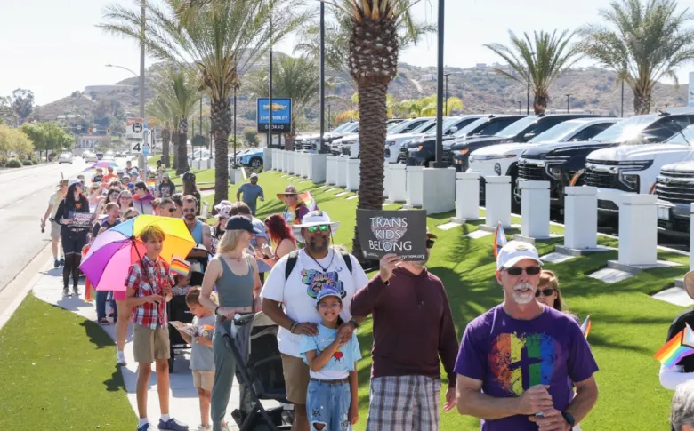 A long line of marchers – one wears a T-shirt with a cross superimposed on a rainbow heart, another holds a rainbow umbrella and another's sign reads "Trans kids belong" – proceeds on a city street past a Chevrolet car dealership.