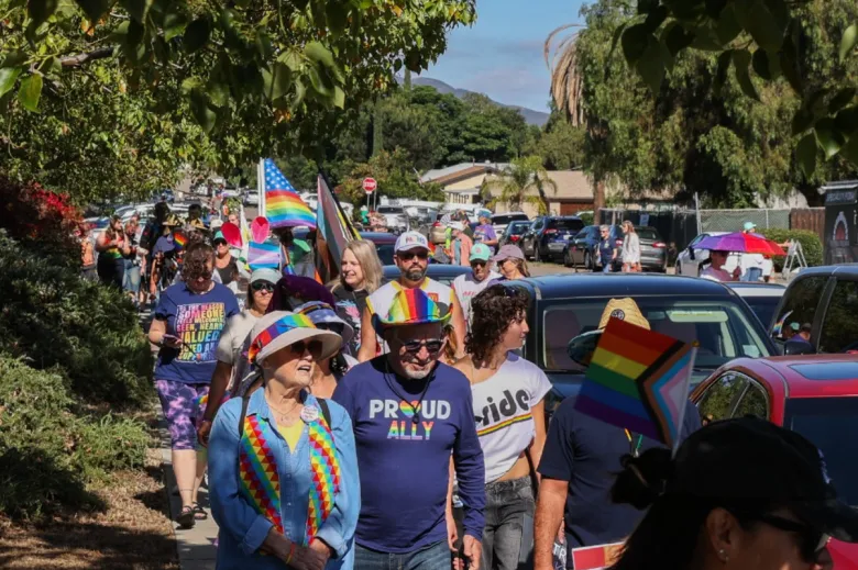 Dozens of people crowd a sidewalk in a long line beside cars and in the shade of several trees. They are wearing shirts that say "Pride" and "Proud ally." Some hold flags in rainbow colors.