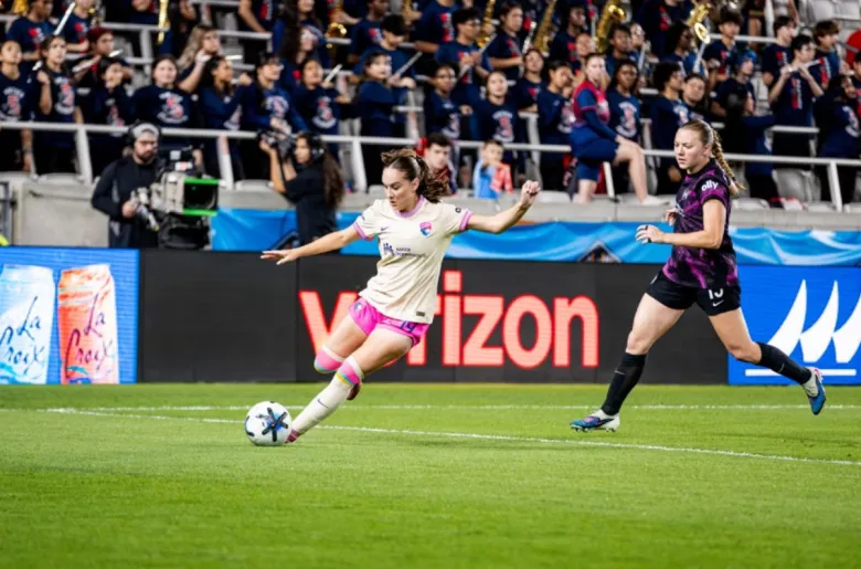 A woman on the soccer pitch with promo banners behind her in front of stands that are filled extends her leg, ready to kick the ball.