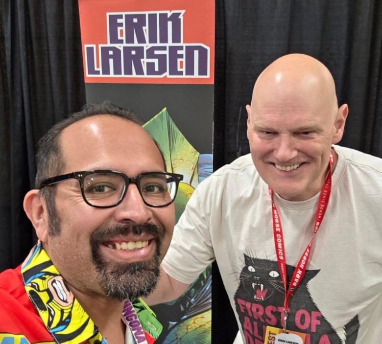 Two men standing in front of a booth with the poster "Erik Larsen."