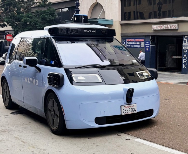 A small car with a camera affixed on top on a city street. On the side of the car and the top is the word "Waymo."