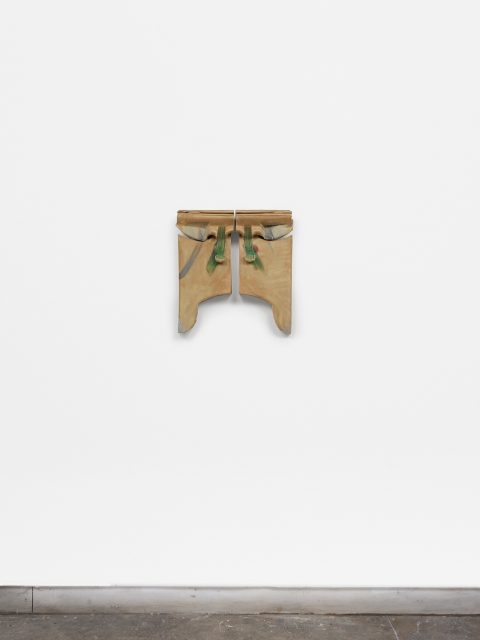 A small, abstract wooden sculpture with green accents is mounted on a white wall above a concrete floor.